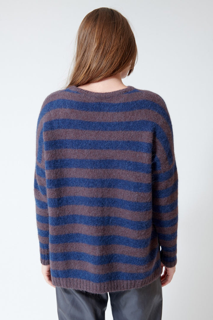 A person shown from the back wearing a loose, fuzzy knit sweater with horizontal stripes in blue and muted taupe-brown, paired with dark gray pants against a plain light background.