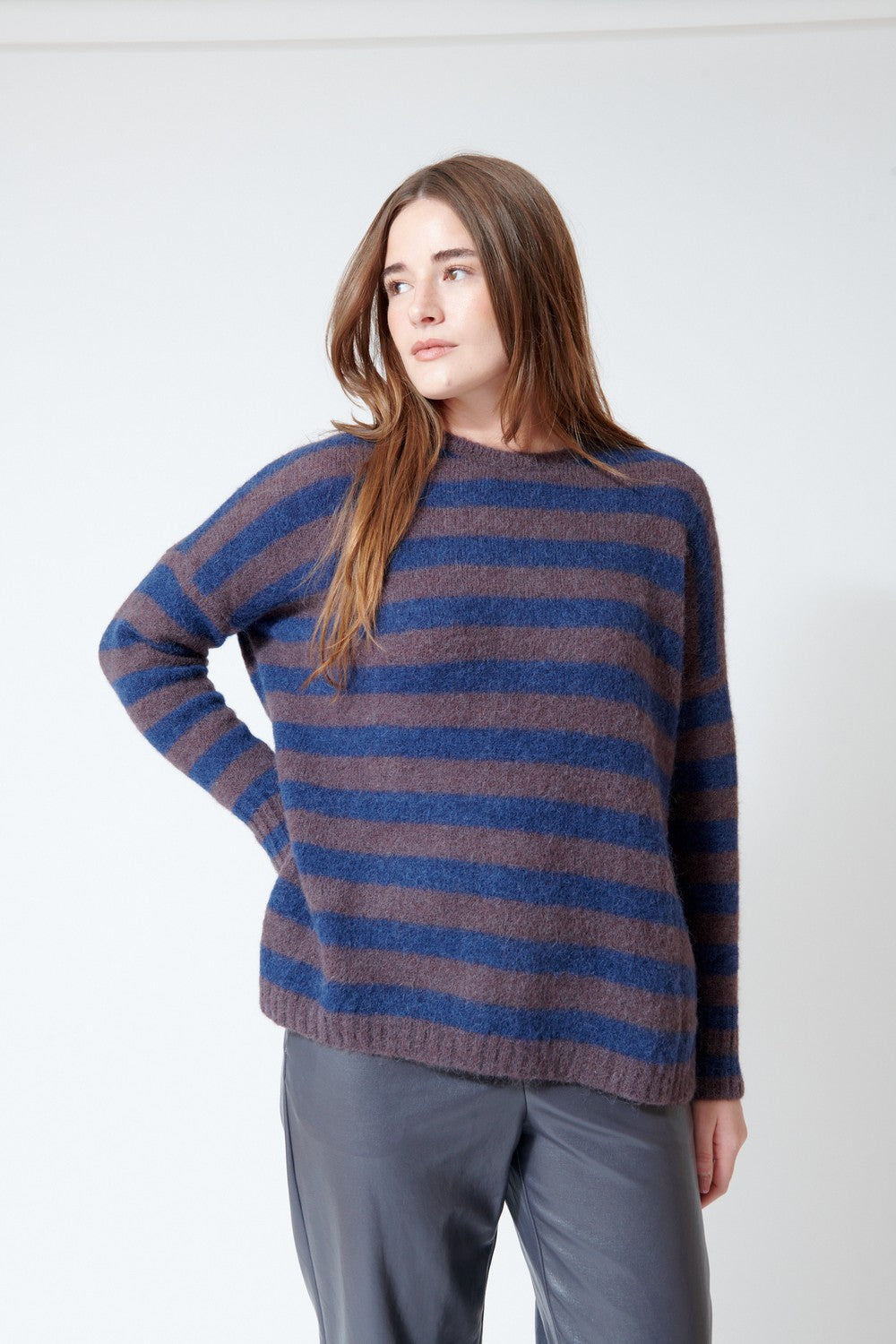 A woman modeling a loose, long-sleeve knit sweater with horizontal stripes in blue and brown, paired with gray pants, against a plain white studio background.