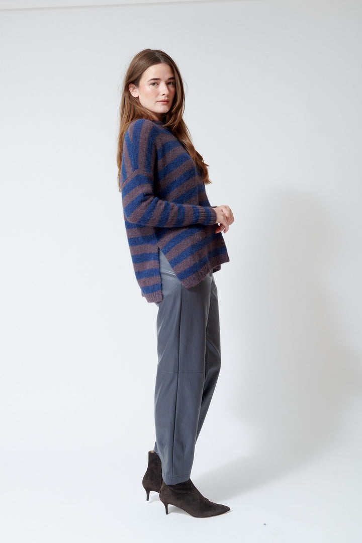A woman modeling a blue and purple striped knit sweater, paired with gray trousers and dark brown pointed-toe ankle boots, standing against a plain white studio background.