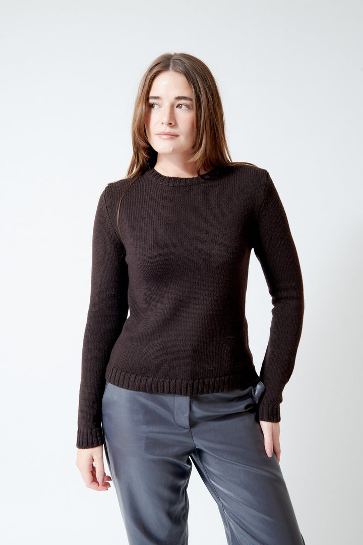 A woman modeling a dark brown ribbed knit crewneck sweater paired with gray pants, photographed against a clean light gray studio background.