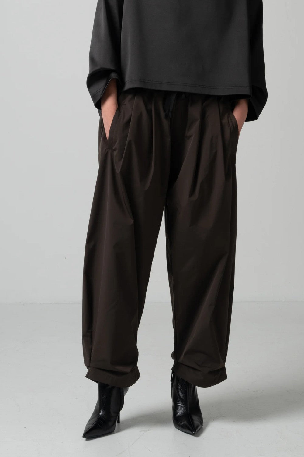 A person wearing loose dark brown drawstring pants with side pockets, paired with a black top and black pointed-toe ankle boots, standing against a light gray studio background.