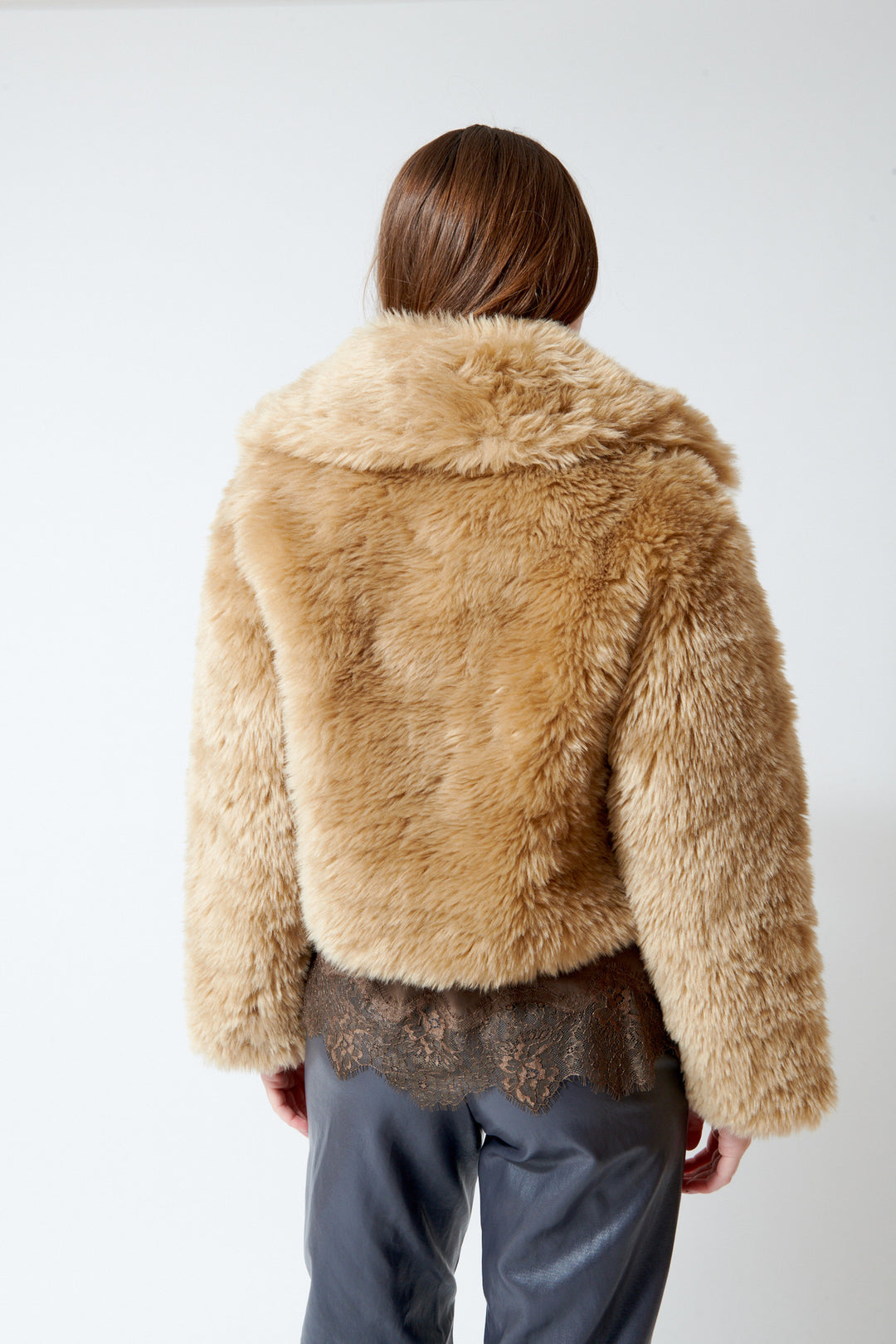 A person shown from the back wearing a  Meimeij cropped, shaggy faux-fur jacket in a warm tan/beige color, paired with dark gray pants and a dark lace hem detail.