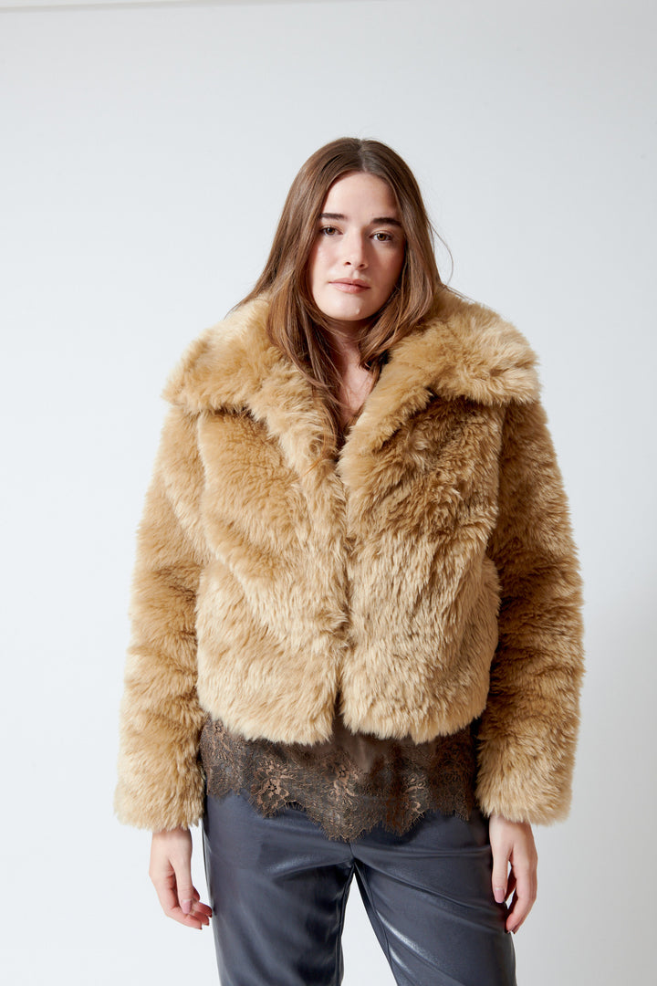 A woman modeling a cropped, shaggy faux-fur jacket in a warm camel tan color by meimeij, sold at Grethen House - front  paired with a dark lace top and dark gray pants, against a plain light gray studio background.