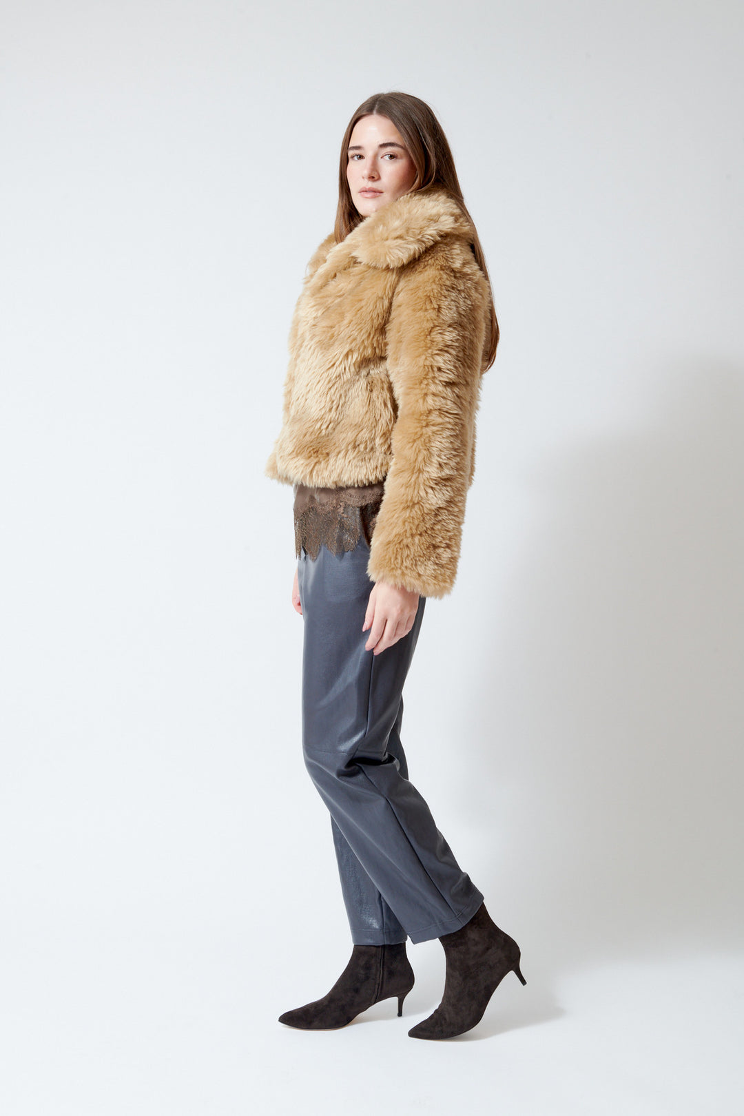 A woman modeling a cropped Meimeij tan faux fur jacket with a wide collar, paired with gray pants and dark brown heeled boots, standing against a plain light gray studio background.
