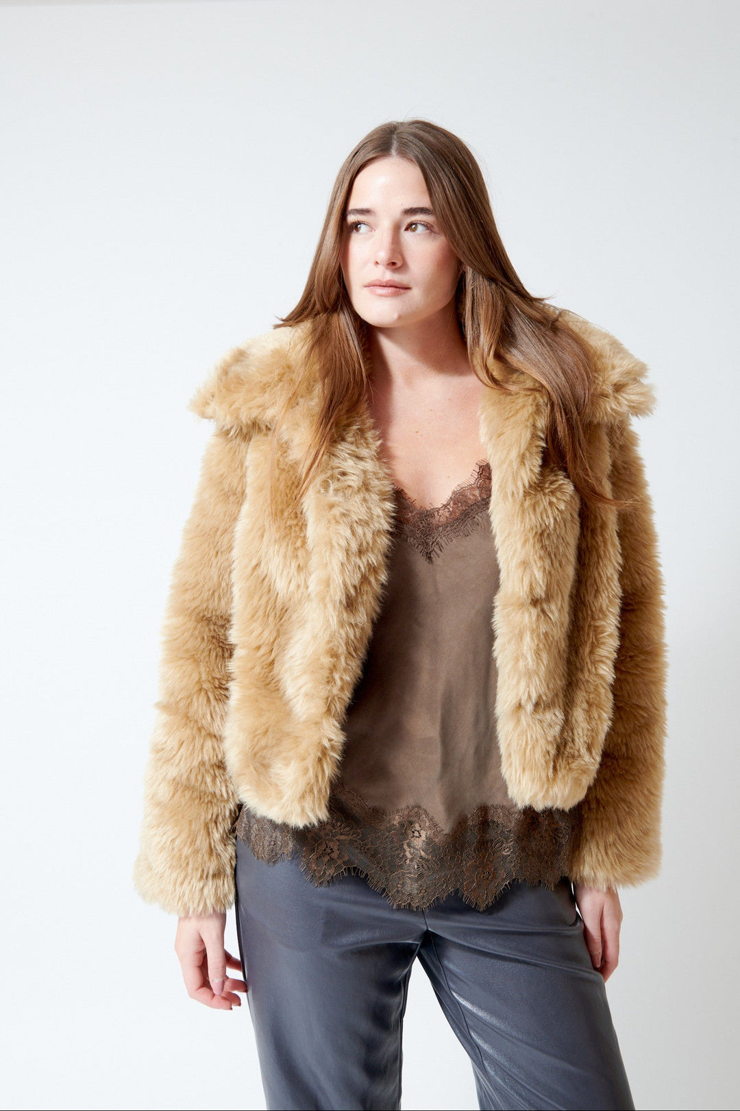 A woman modeling a cropped Meimeij, fluffy faux-fur jacket in a warm tan/beige color, worn over a sheer brown lace camisole and dark gray trousers, against a plain white studio background.