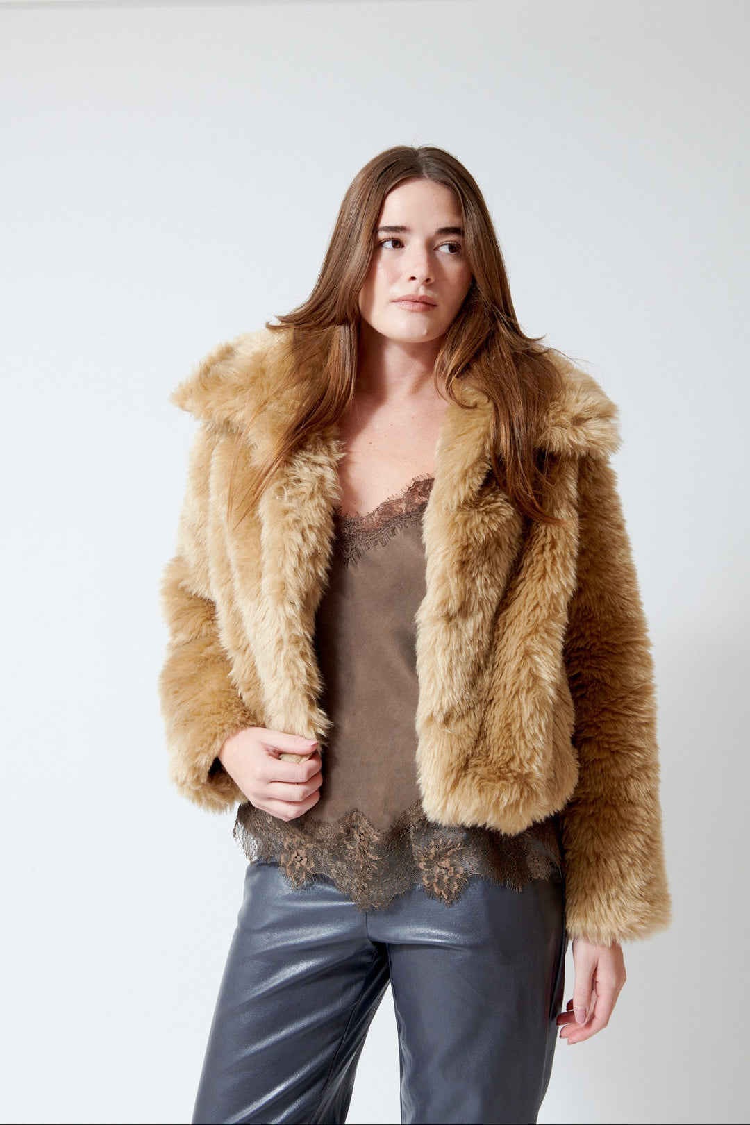 A woman modeling a cropped, plush faux-fur jacket in a warm camel brown color, worn over a brown lace-trim camisole and dark gray pants, against a plain white studio background.