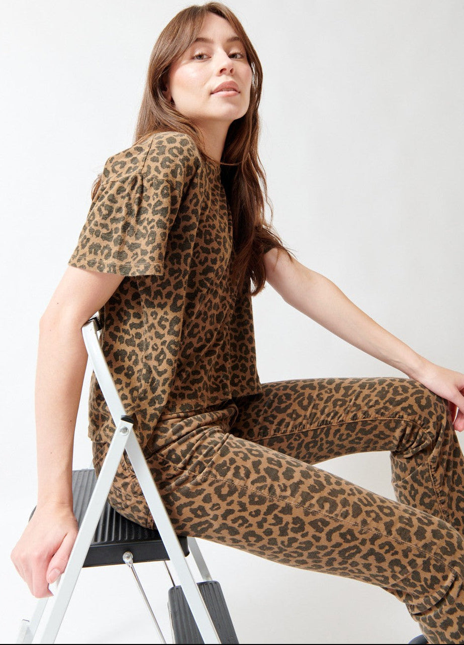 A woman wearing a matching leopard-print short-sleeve top and pants set, seated on a white step ladder against a plain white studio background.