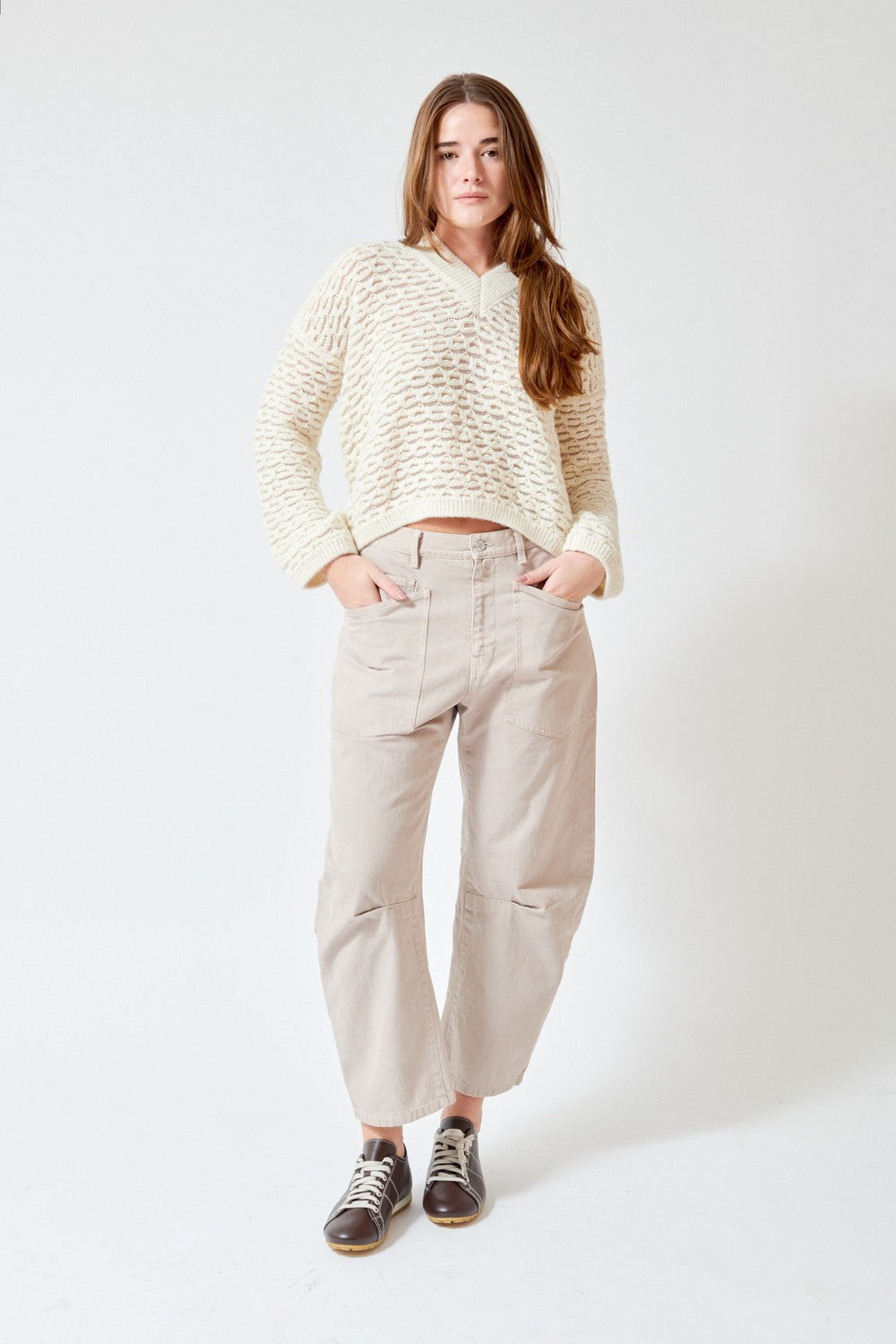 A woman stands against a plain white studio background wearing a cream-colored textured knit sweater, beige wide-leg cropped pants, and dark brown sneakers.