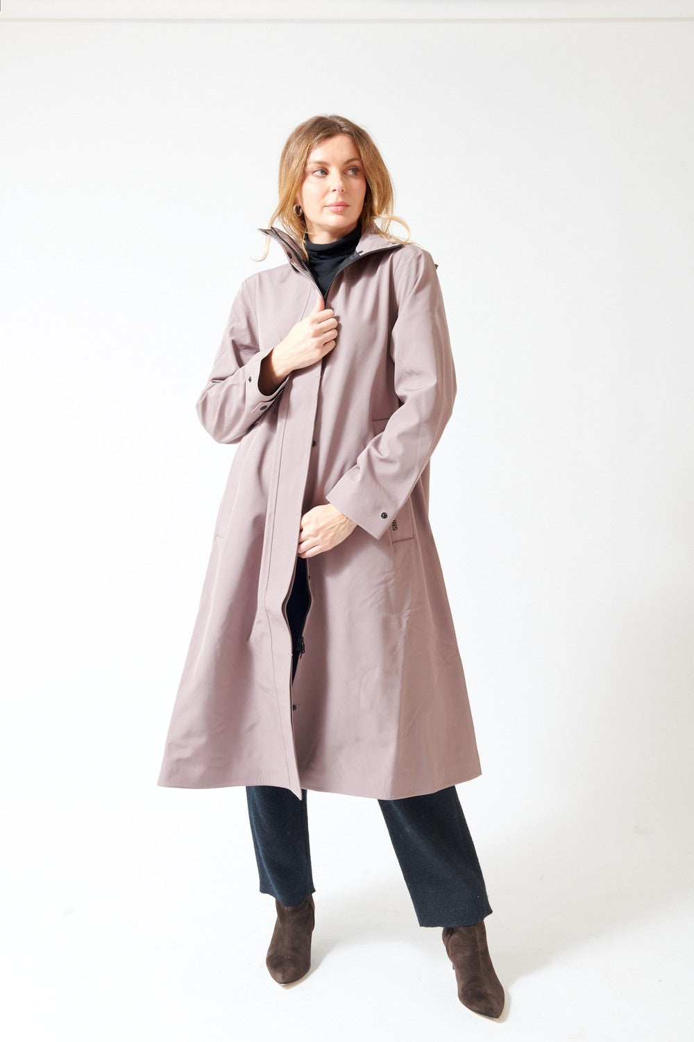 A woman modeling a long, mid-length dusty mauve raincoat over a black turtleneck, dark trousers, and brown ankle boots, standing against a plain white studio background.