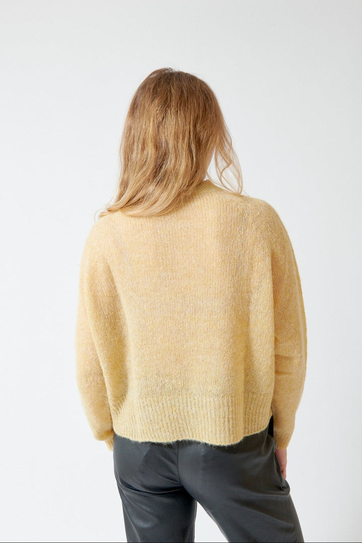 A woman viewed from behind wearing a fuzzy, cropped pale yellow knit sweater and dark gray pants, standing against a plain white wall.