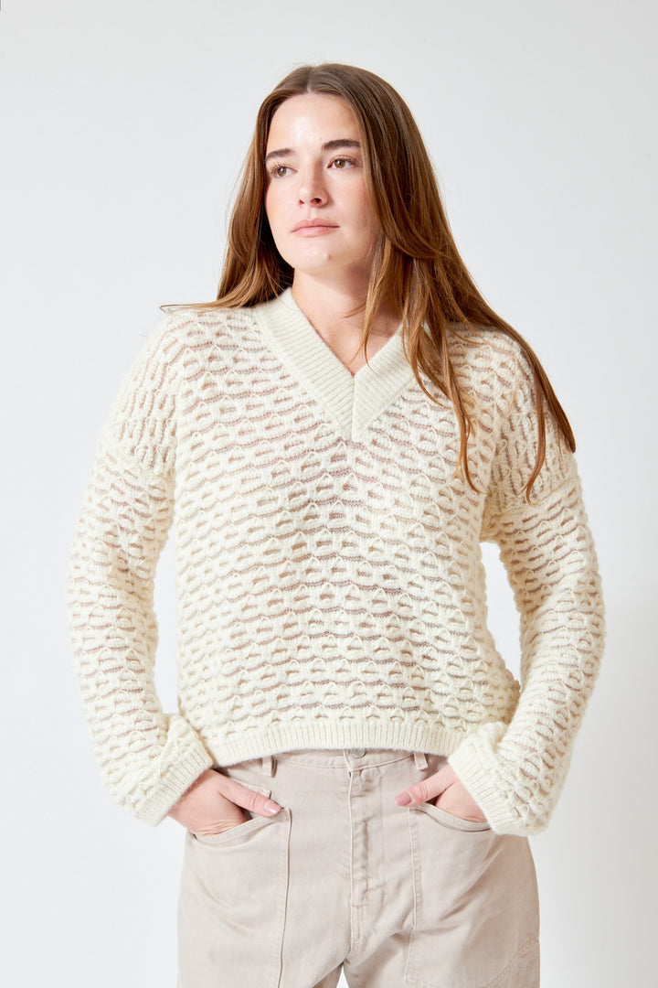 A woman wearing a cream-colored textured knit sweater with a V-neck and beige pants, standing against a light gray studio background.