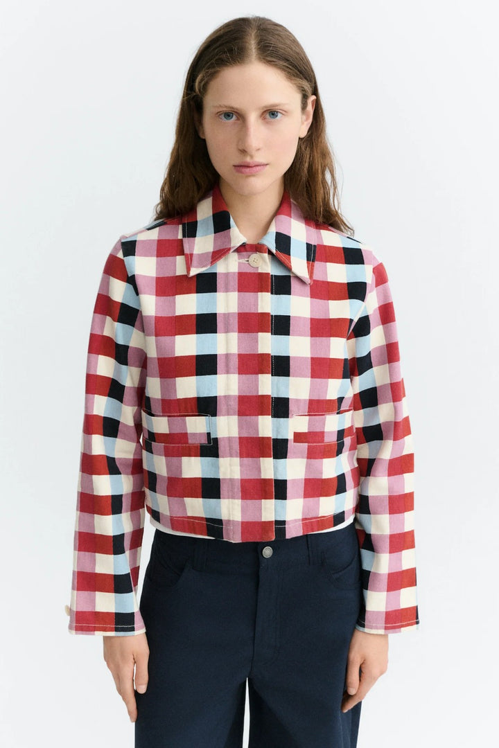 A model wearing a cropped, long-sleeve plaid button-up shirt with a red, white, black, and light blue checkered pattern, paired with dark navy trousers, against a plain white studio background.