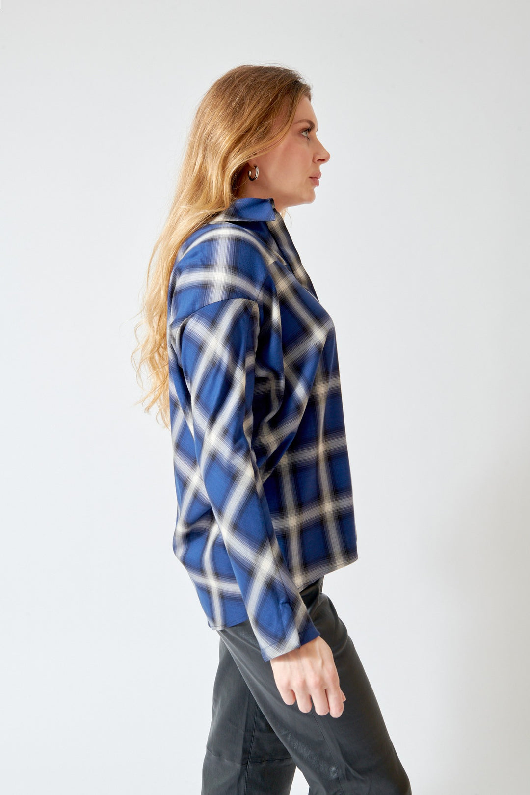 A woman in profile wearing a blue, white, and black plaid button-up shirt and dark gray jeans, standing against a plain light gray background.