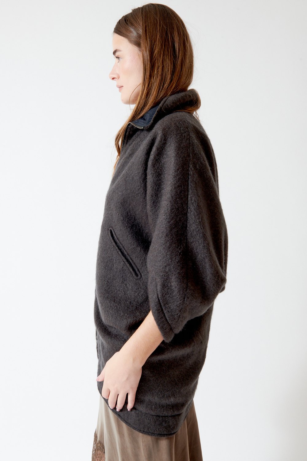 A woman shown in profile wearing a dark charcoal gray fuzzy half-zip pullover jacket with a high collar and a zippered pocket, paired with a brown skirt, against a plain white studio background.