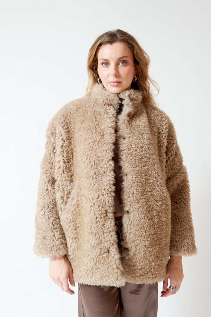 A woman wearing a beige curly teddy coat with a high collar, styled over a brown top and brown pants, photographed against a plain white studio background.