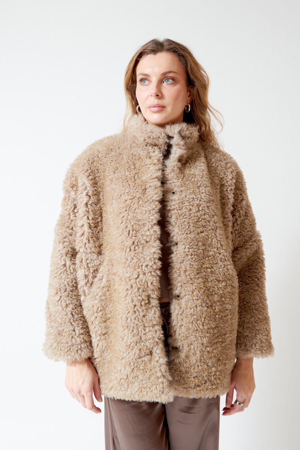 A woman wearing a beige curly-textured shearling-style jacket with a high collar and front buttons, paired with brown pants, photographed against a plain light gray studio background.
